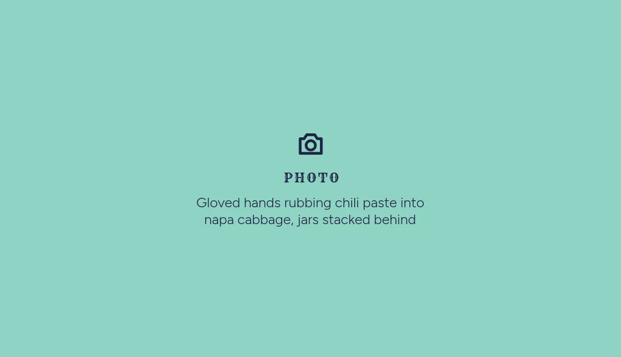Photo placeholder: Gloved hands rubbing chili paste into napa cabbage, jars stacked behind